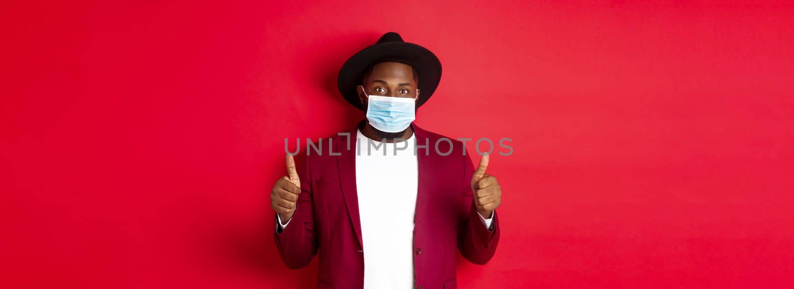 Covid-19 and fashion concept. Stylish african american man in hat and blazer, wearing face mask and showing thumb up, standing over red background.