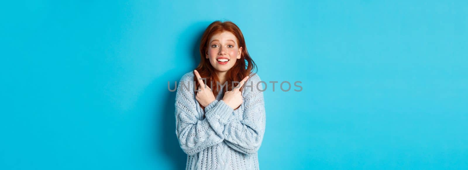 Excited redhead girl pointing fingers sideways, showing two choices and looking tempted at camera, standing against blue background.