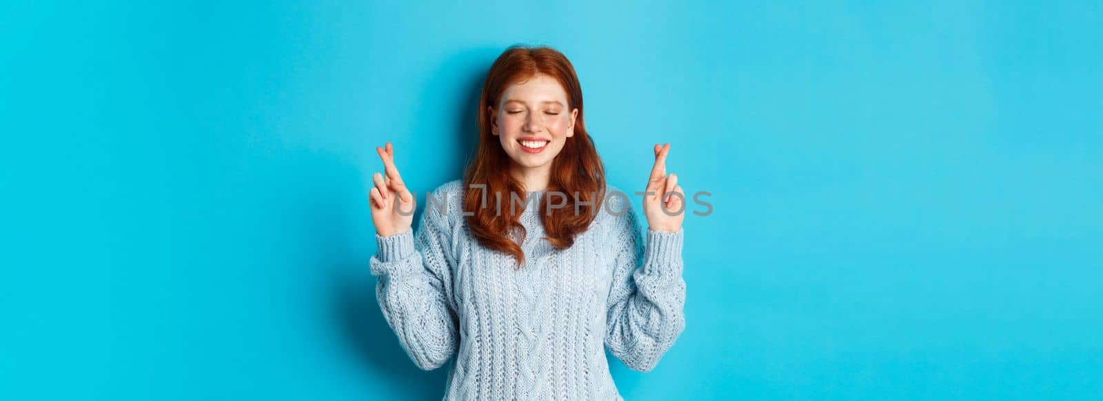 Hopeful redhead girl making a wish, cross fingers for good luck, smiling and anticipating good news or positive result, standing against blue background.