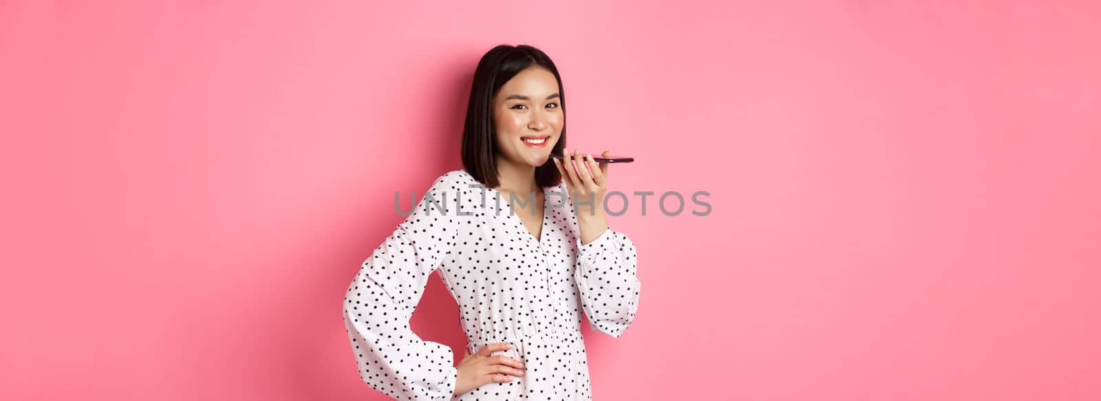 Beautiful korean woman talking on speakerphone, recording voice message and smiling happy, standing over pink background.