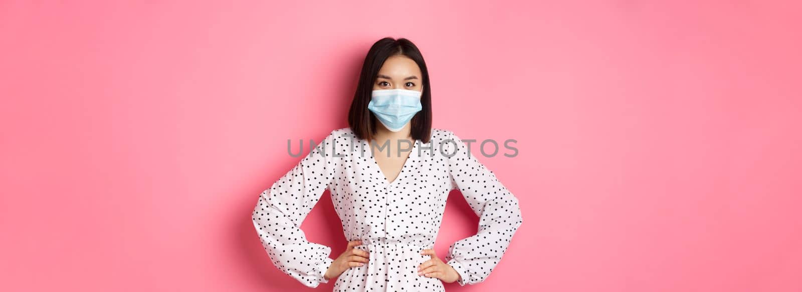 Covid-19, quarantine and lifestyle concept. Cute asian woman wearing medical mask and dress, protect herself from coronavirus, standing over pink background.