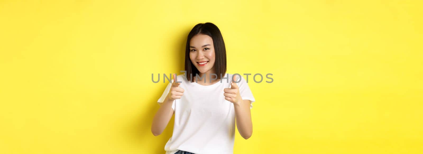 Pretty young asian woman in white t-shirt, pointing fingers at camera and smiling, praising or choosing you, say congrats, standing over yellow background by Benzoix