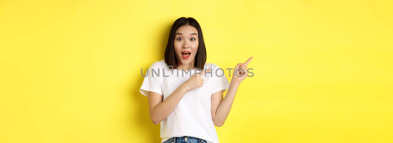 Beauty and fashion concept. Beautiful asian woman in white t-shirt pointing fingers left, standing over yellow background by Benzoix
