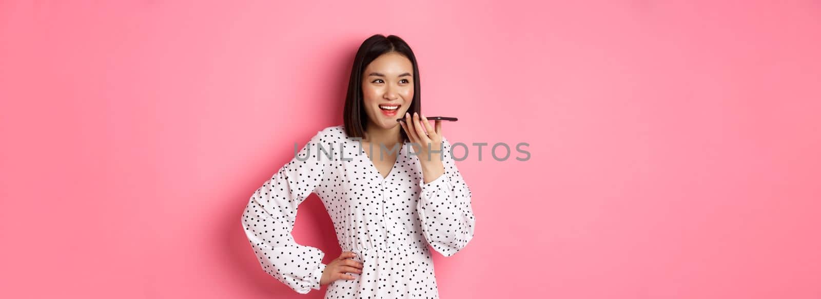 Beautiful korean woman talking on speakerphone, recording voice message and smiling happy, standing over pink background.