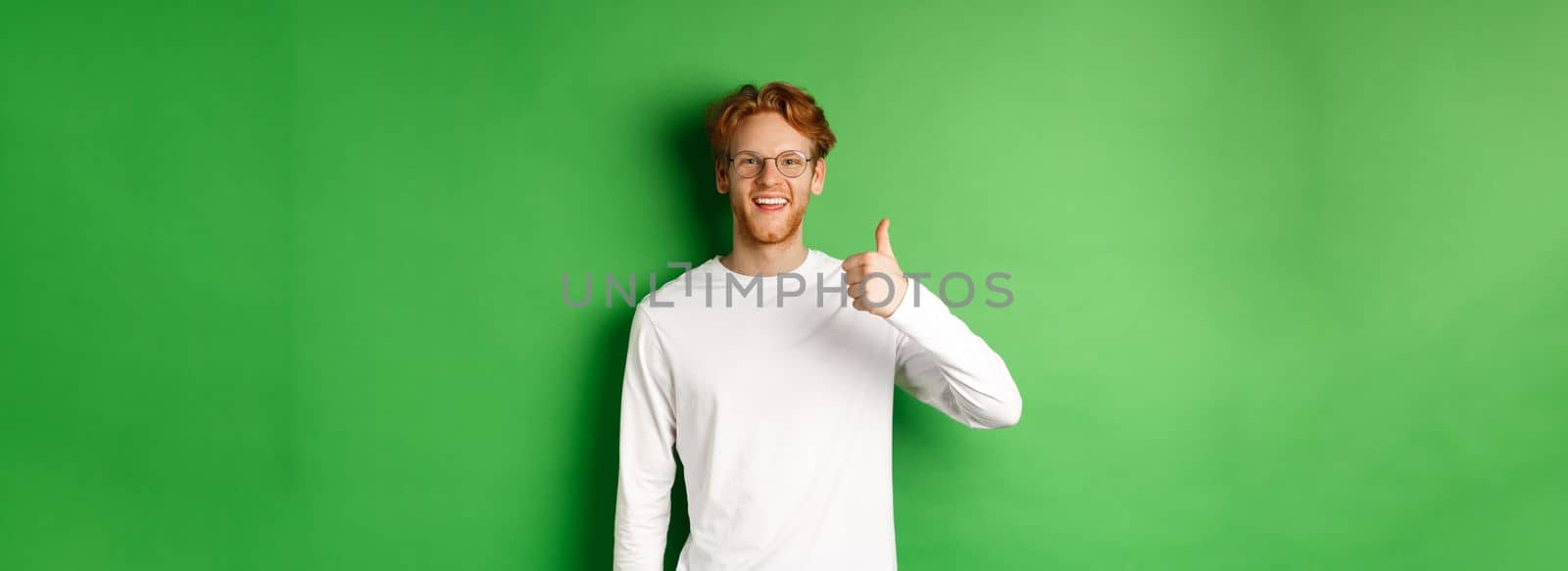 Handsome young man in glasses with red hair, showing thumbs-up and smiling satisfied, praising good eyewear store, standing over green background.