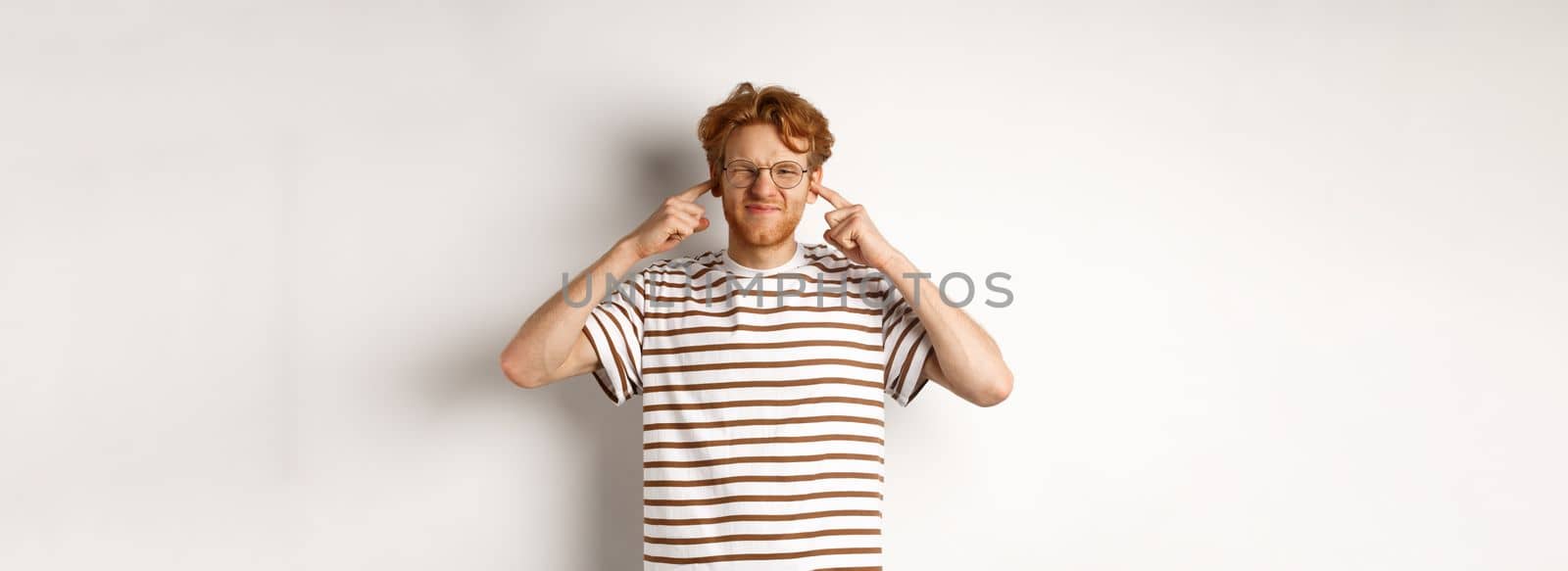 Annoyed young man complaining on loud noise, shut ears and grimacing from discomfort, standing over white background.