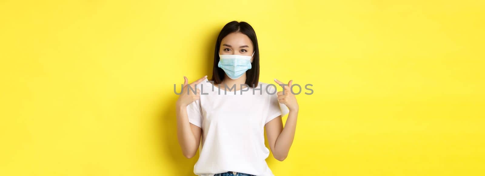 Covid-19, quarantine and social distancing concept. Young asian woman in white t-shirt, pointing at her medical mask from coronavirus, yellow background.