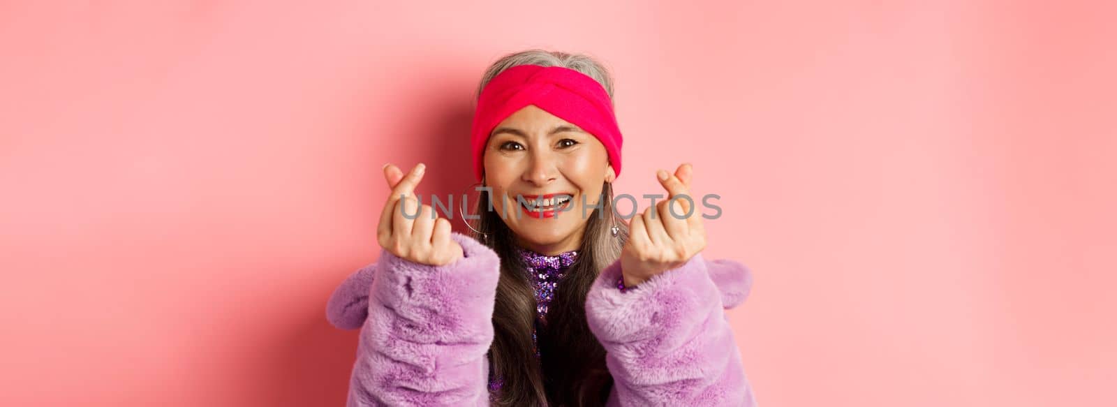 Fashion. Beautiful and fashionable asian senior woman smiling, showing korean heart gesture and looking happy, standing over pink background.