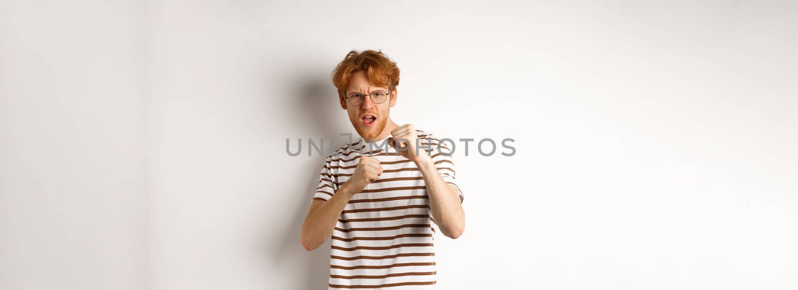 Funny young man with red hair raising fists for fight, shadow boxing and looking serious at camera, standing over white background.