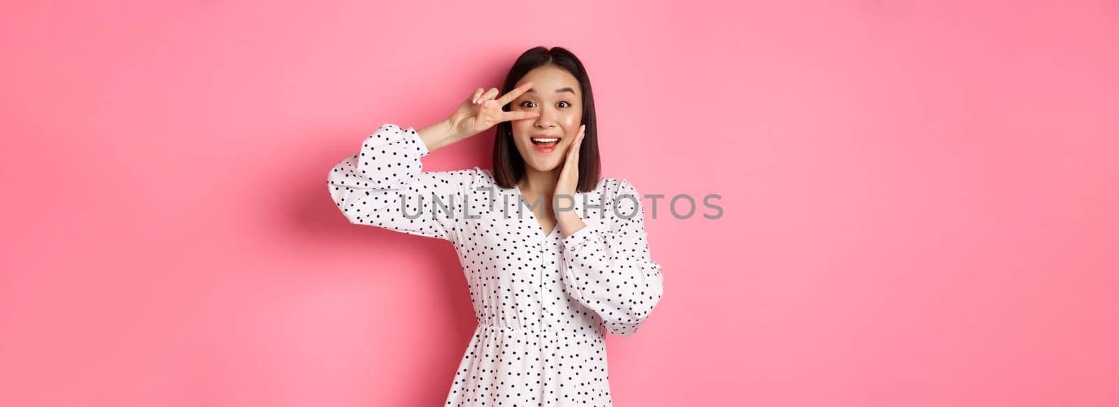 Beautiful and feminine asian woman showing kawaii sign on eye and looking amazed at camera, standing in dress over pink background.