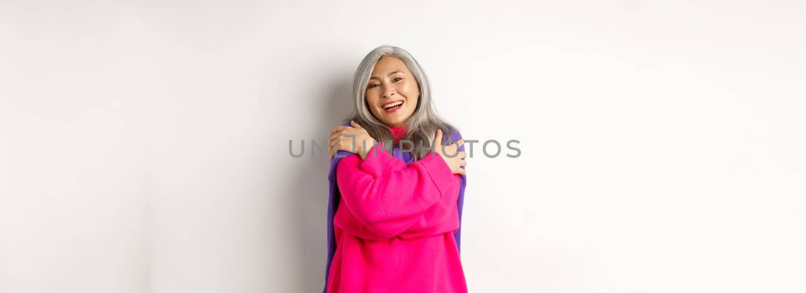 Valentines day and holidays concept. Lovely asian senior woman in pink sweater hugging herself with eyes closed, smiling, standing over white background by Benzoix