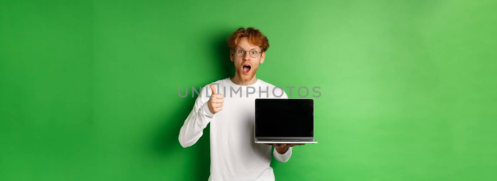 Impressed caucasian man with red hair and beard, showing blank laptop screen and thumb-up, looking amazed at camera, praising something online, green background.
