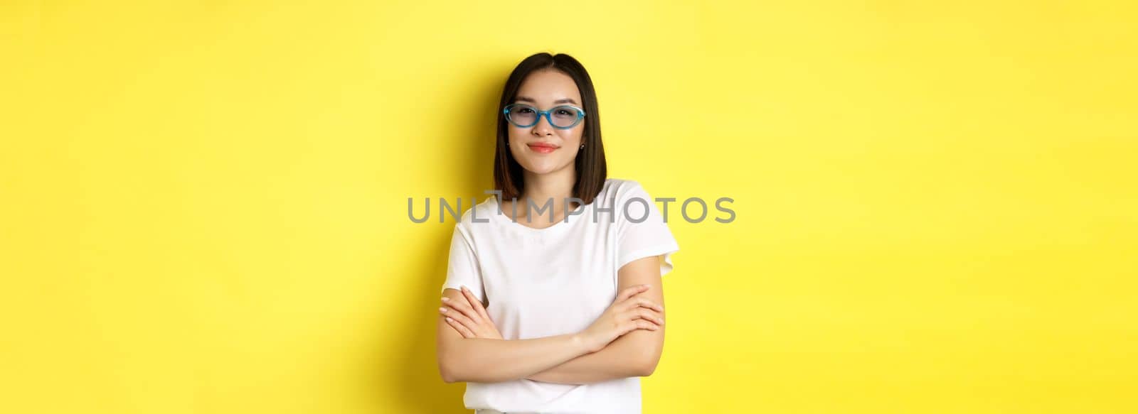 Fashion and lifestyle concept. Sassy and confident asian woman in trendy sunglasses looking self-assured at camera, standing over yellow background.