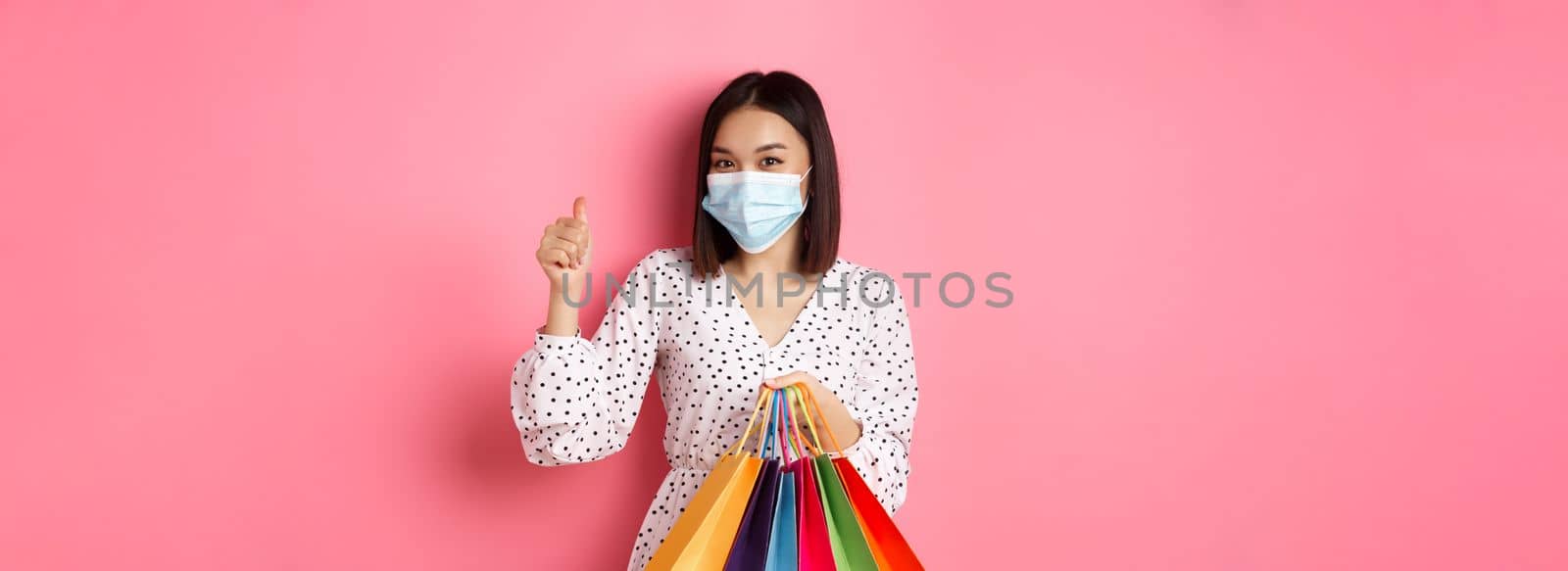 Covid-19, pandemic and lifestyle concept. Beautiful asian woman shopping in malls in medical mask, holding bags and showing thumb-up, standing over pink background.