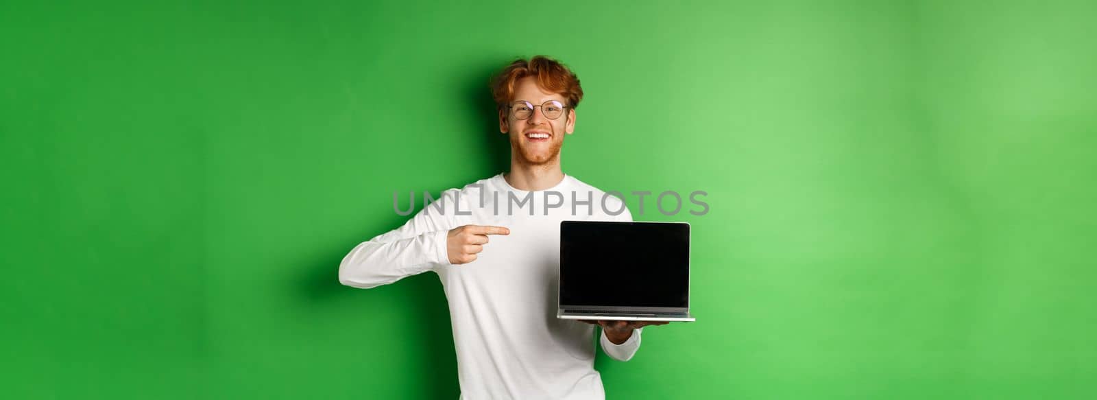 Happy redhead guy in glasses and white long-sleeve t-shirt, pointing finger at blank laptop screen and smiling, standing over green background.