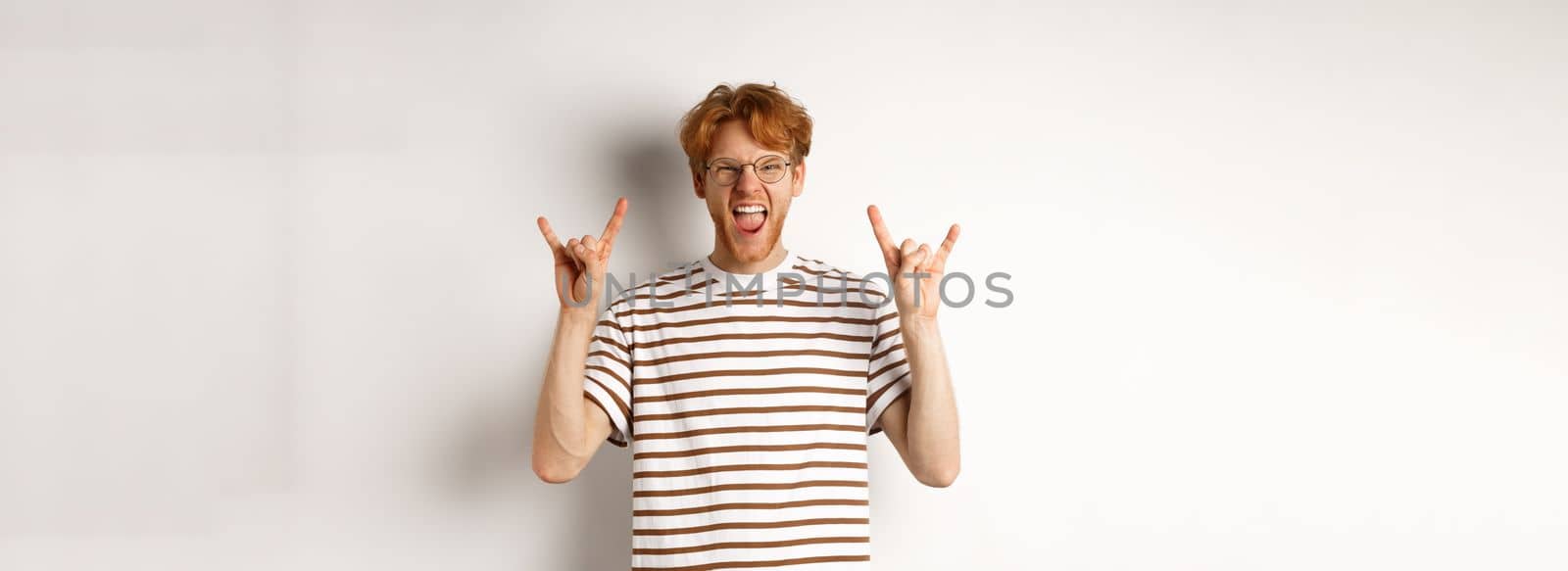 Funny and happy redhead man having fun, showing rock-n-roll horn and sticking tongue, enjoying party, standing over white background by Benzoix