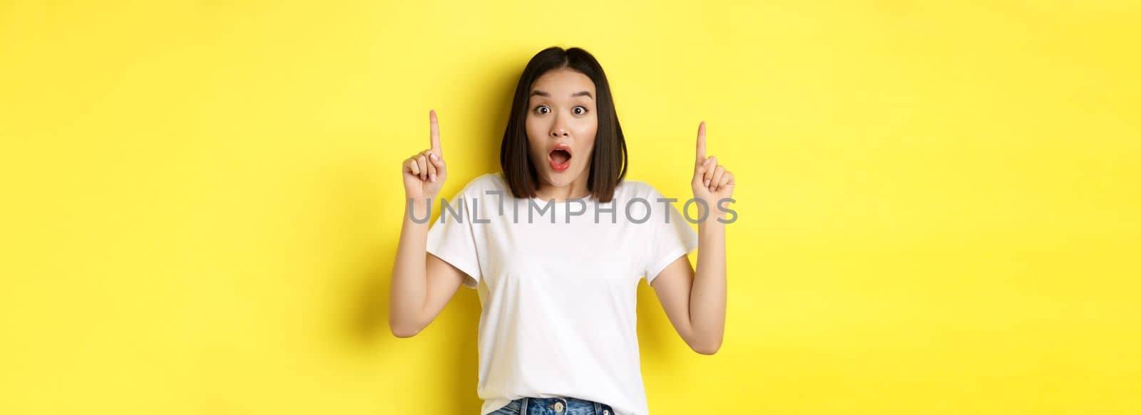 Beauty and fashion concept. Beautiful asian woman in white t-shirt pointing fingers up, standing over yellow background.