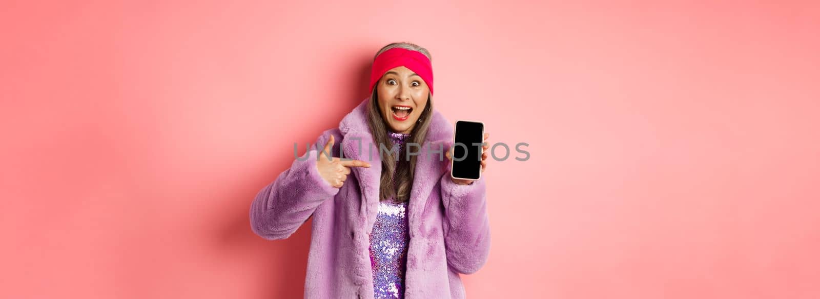 Online shopping and fashion concept. Stylish asian senior woman in faux fur coat pointing finger at blank smartphone screen, smiling happy, showing application on phone.