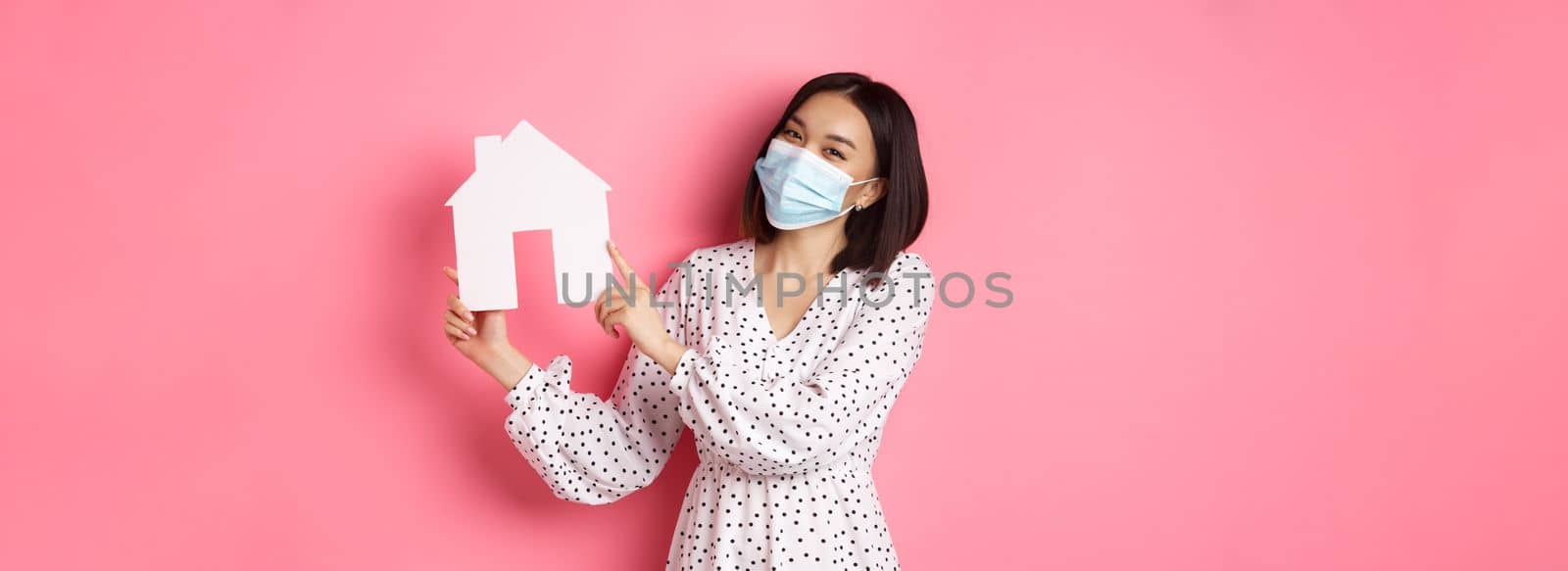 Covid-19, real estate and lifestyle concept. Cute asian woman in face mask selling houses, showing model of home and looking at camera, standing over pink background by Benzoix