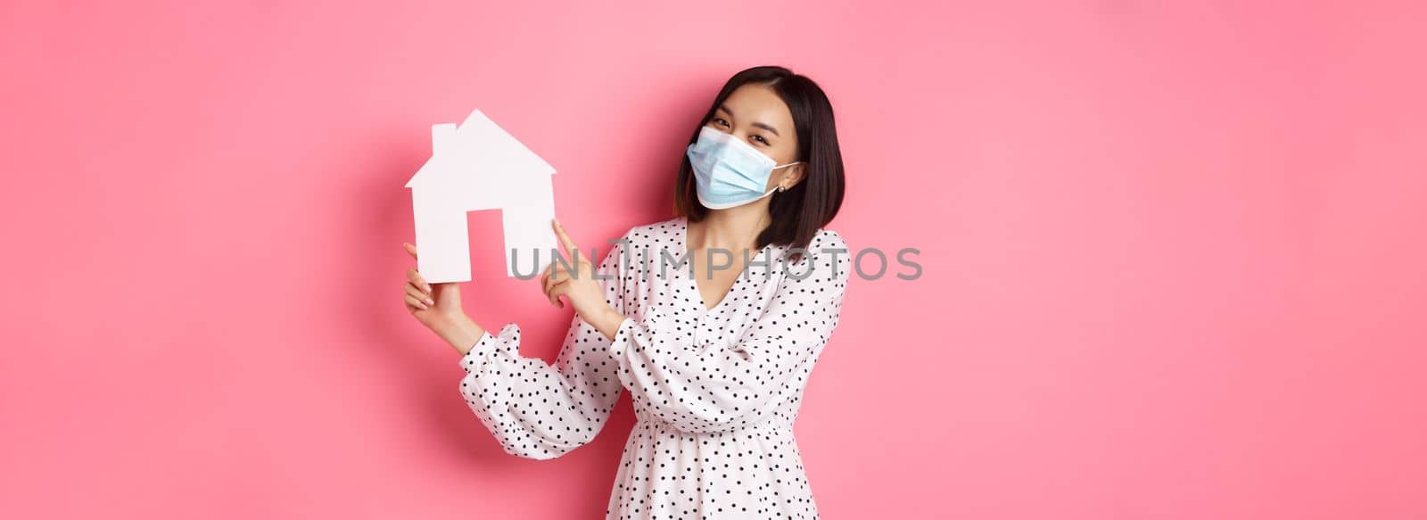 Covid-19, real estate and lifestyle concept. Cute asian woman in face mask selling houses, showing model of home and looking at camera, standing over pink background.