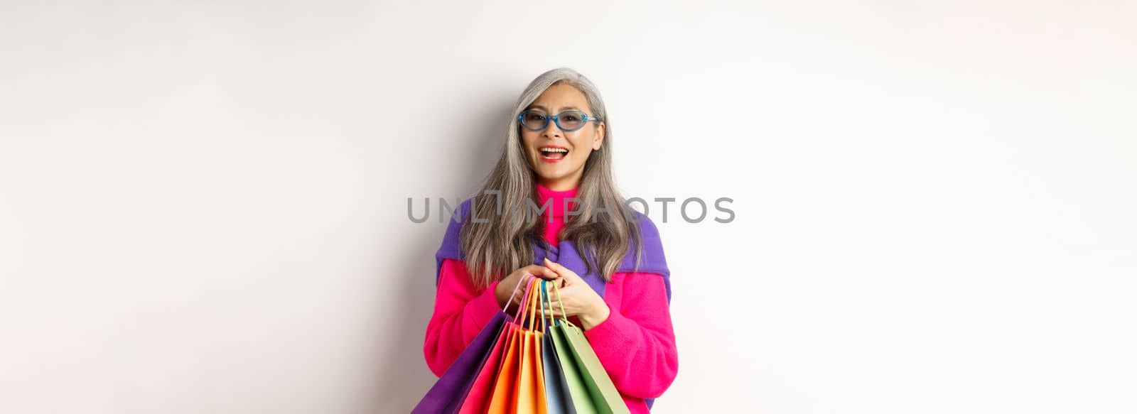 Stylish senior asian woman in sunglasses going shopping on holiday sale, holding paper bags and smiling, standing over white background.