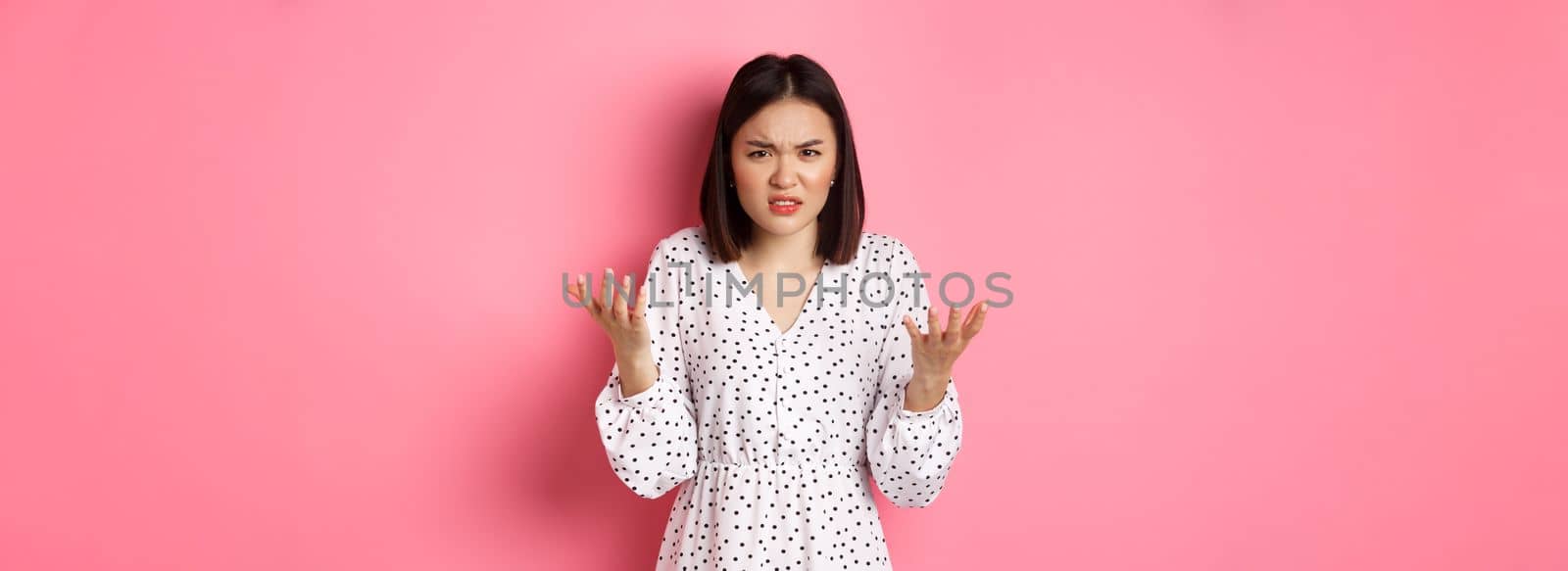 Angry and confused asian woman pointing hands at camera and grimacing furious, standing annoyed against pink background.