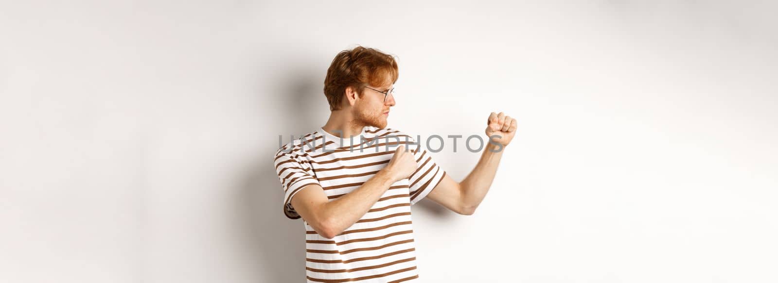 Funny young man with red hair raising fists for fight, shadow boxing and looking serious at left side, standing over white background.
