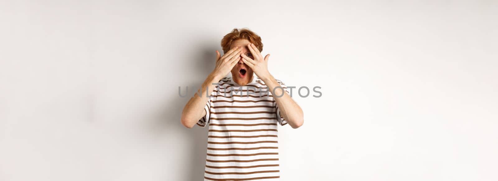 Shocked and embarrassed redhead guy covering eyes, peeking through fingers and gasping, standing over white background.