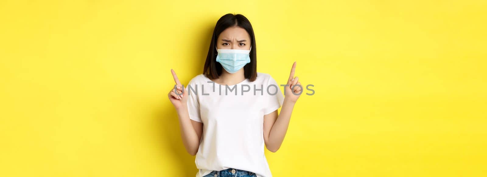 Covid-19, pandemic and social distancing concept. Disappointed asian girl in medical mask, frowning upset and pointing fingers up at logo, standing over yellow background.