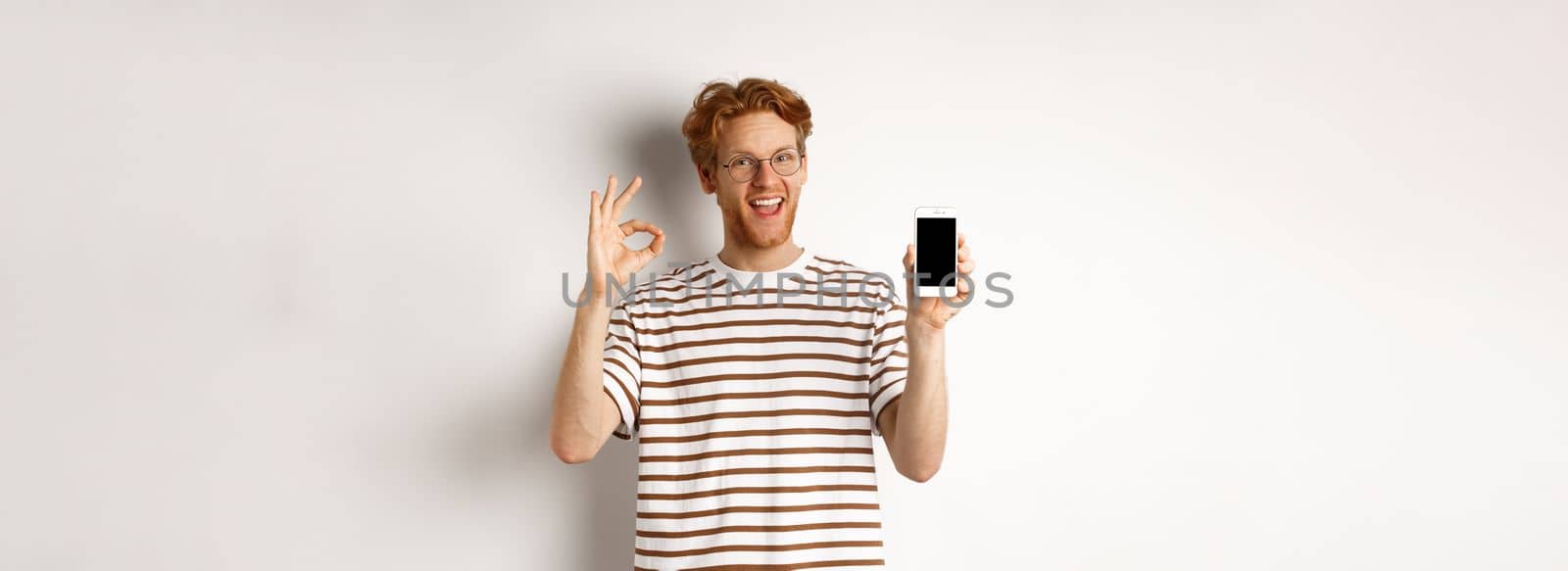 Technology and e-commerce concept. Young man with red hair showing okay sign and blank smartphone screen, praising awesome app, standing over white background by Benzoix