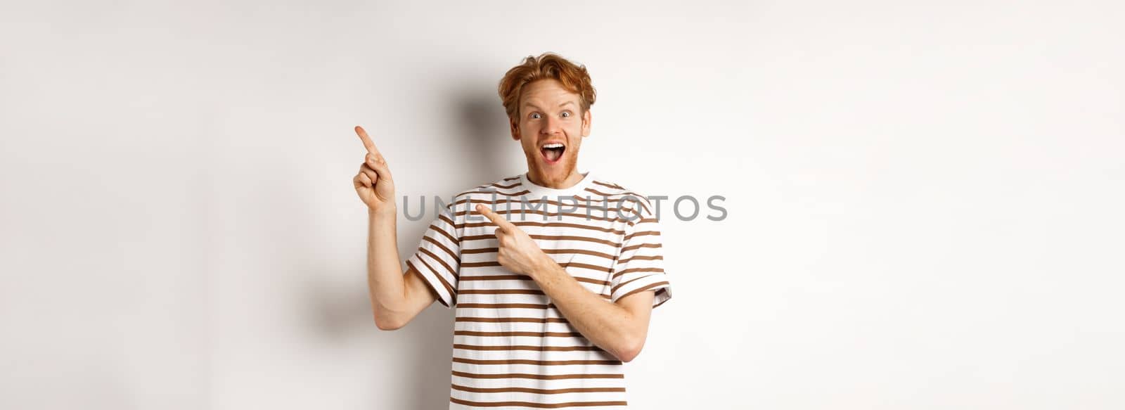 Excited redhead guy in striped t-shirt checking out special deal, pointing fingers right and smiling at camera, white background.