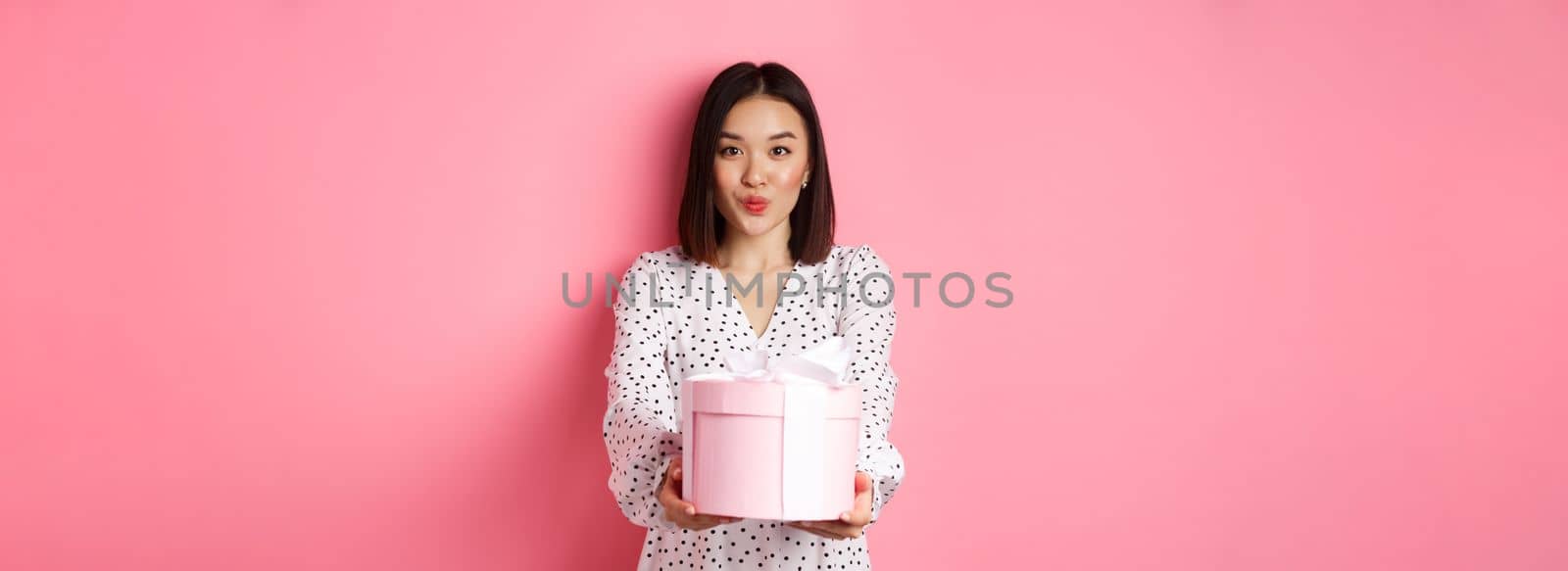Cute asian girlfriend congratulate with valentines day, giving cute romantic gift in box, pucker lips for kiss, standing over pink background by Benzoix