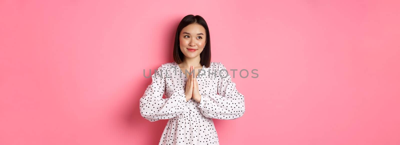 Beautiful angelic asian woman smiling, holding hands in pray and looking left at copy space with innocent cute gaze, standing over pink background by Benzoix