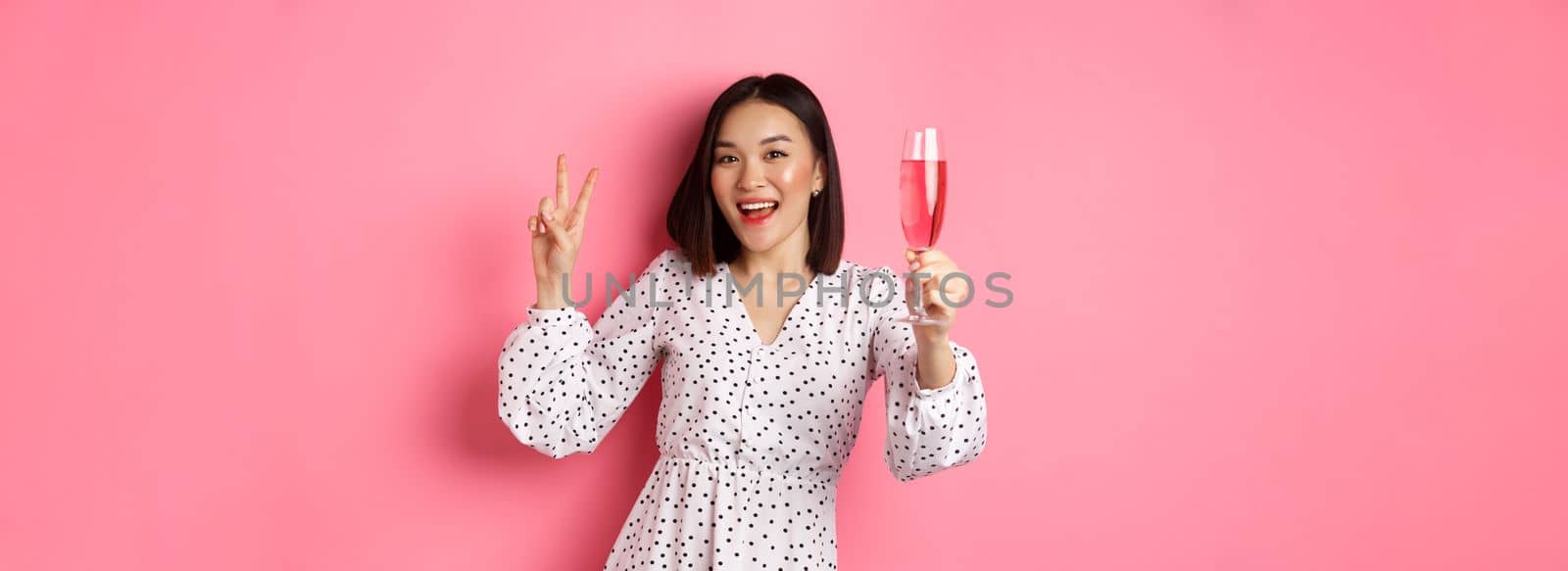 Cute asian female model drinking champagne, celebrating on party and showing peace sign, smiling happy at camera, standing over pink background.