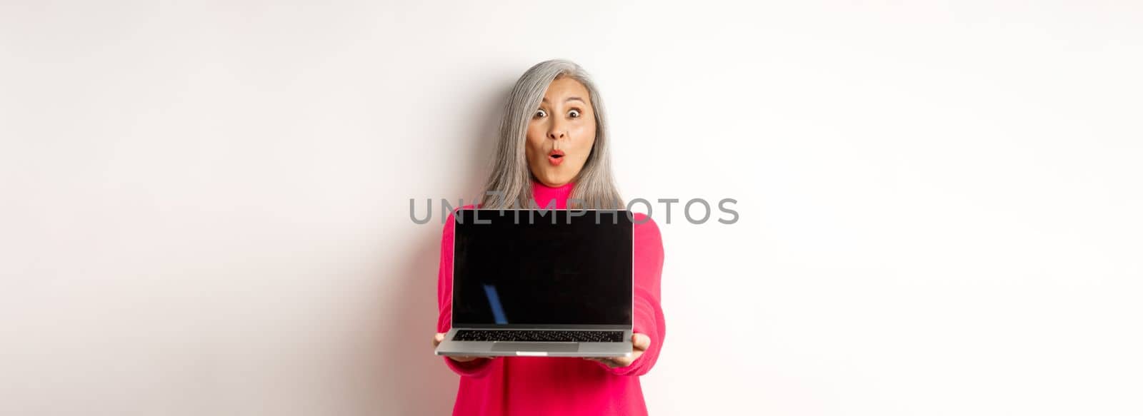 E-commerce concept. Impressed asian senior woman showing blank laptop screen and staring amazed at camera, standing over white background.