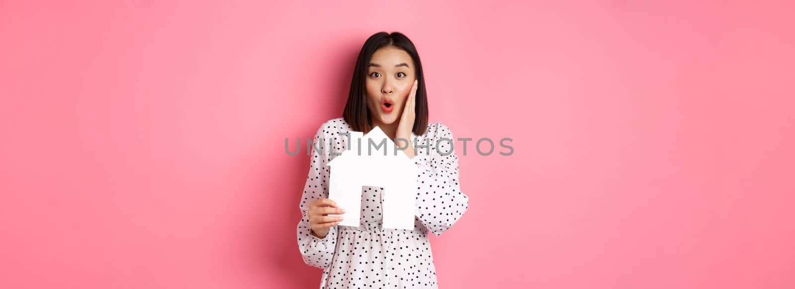 Real estate. Surprised and amazed asian woman staring at camera, saying wow and showing paper house model, standing over pink background.