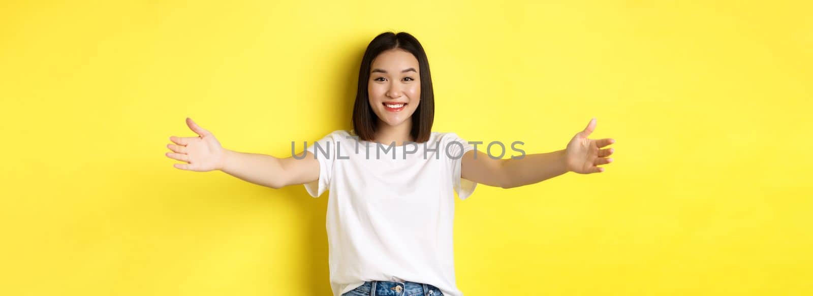Beauty and fashion concept. Friendly asian woman spread out hands and smiling, waiting for hugs, inviting you, welcome someone and looking happy, standing over yellow background by Benzoix