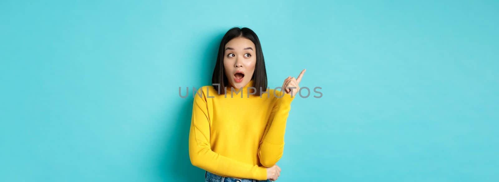 Shopping concept. Impressed asian girl in yellow pullover, pointing and looking left amazed, showing logo banner, standing over blue background.