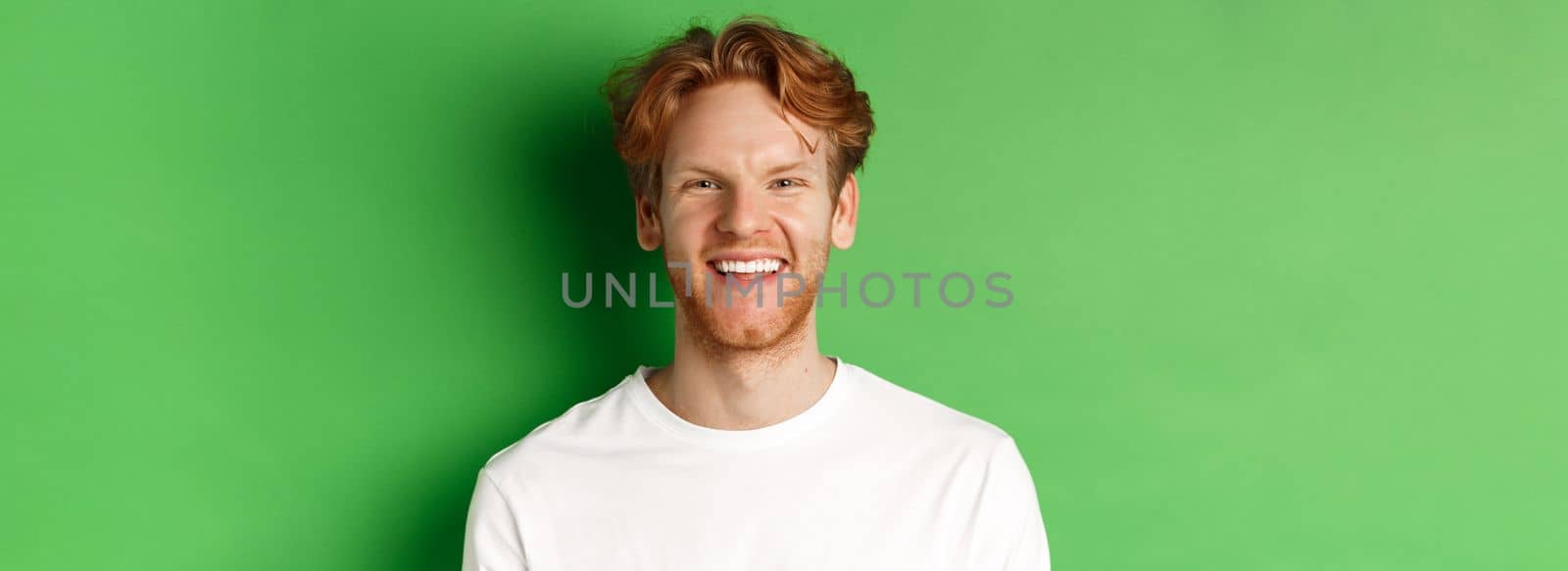 Emotions and fashion concept. Happy young man with red hair and beard, smiling and laughing at camera, standing over green background.