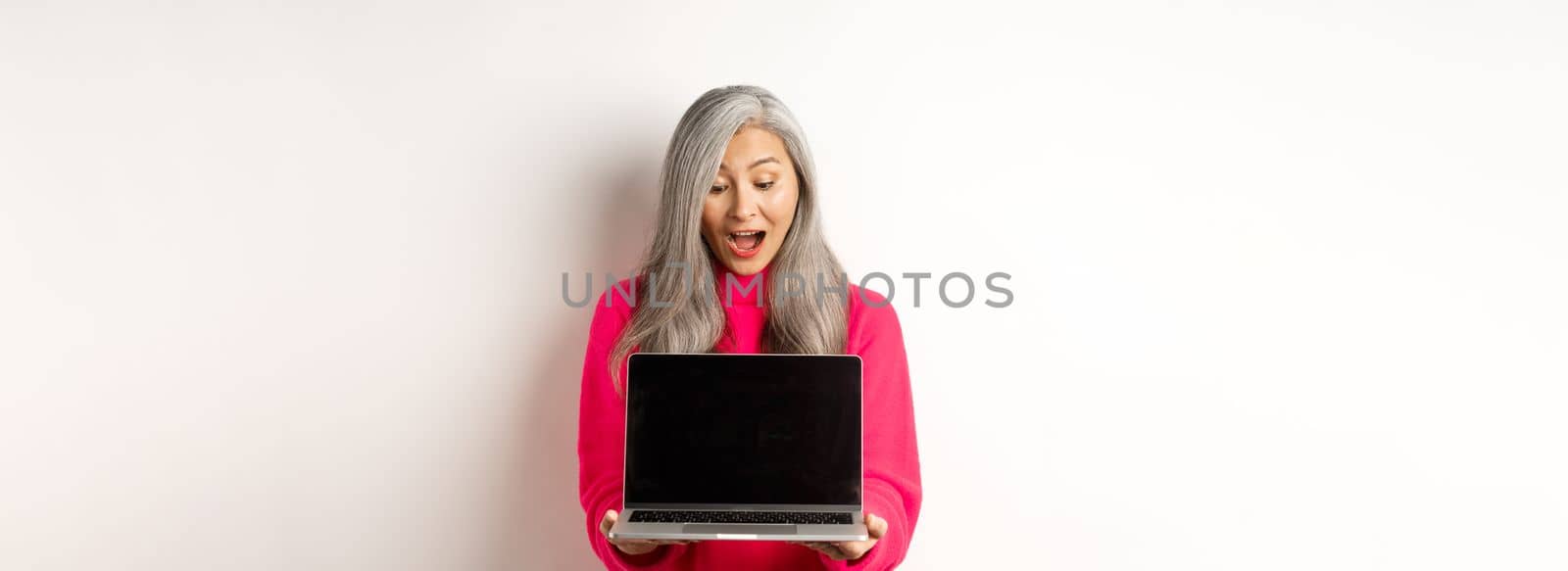 E-commerce concept. Amazed asian grandmother with grey hair, checking out promo online, showing laptop black screen, standing over white background.