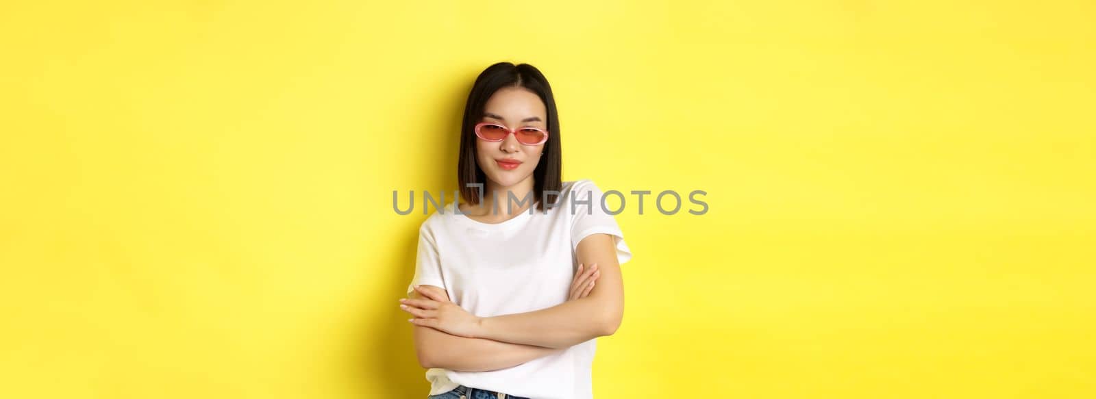 Fashion and lifestyle concept. Sassy and confident asian woman in trendy sunglasses looking self-assured at camera, standing over yellow background by Benzoix