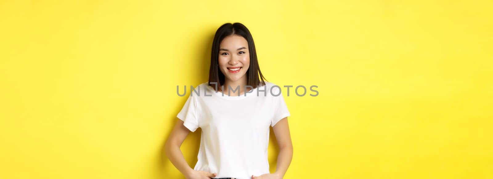 Beauty and fashion concept. Stylish asian girl in white t-shirt, smiling happy and standing over yellow background.