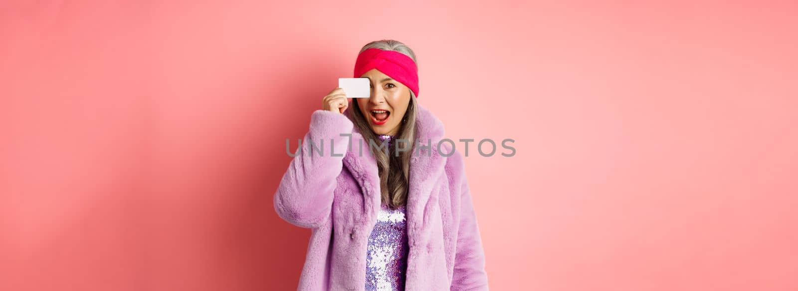Shopping and fashion concept. Beautiful asian middle-aged woman showing plastic credit card on face and looking excited at camera, standing over pink background.