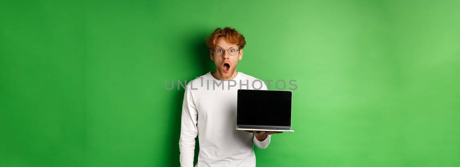 Impressed redhead guy in glasses drop jaw after seeing online promo, showing laptop screen and staring at camera with disbelief, standing over green background by Benzoix