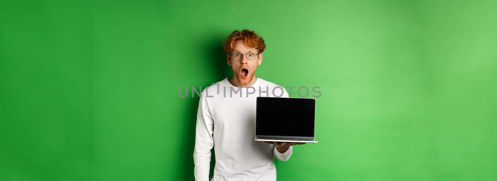 Impressed redhead guy in glasses drop jaw after seeing online promo, showing laptop screen and staring at camera with disbelief, standing over green background.