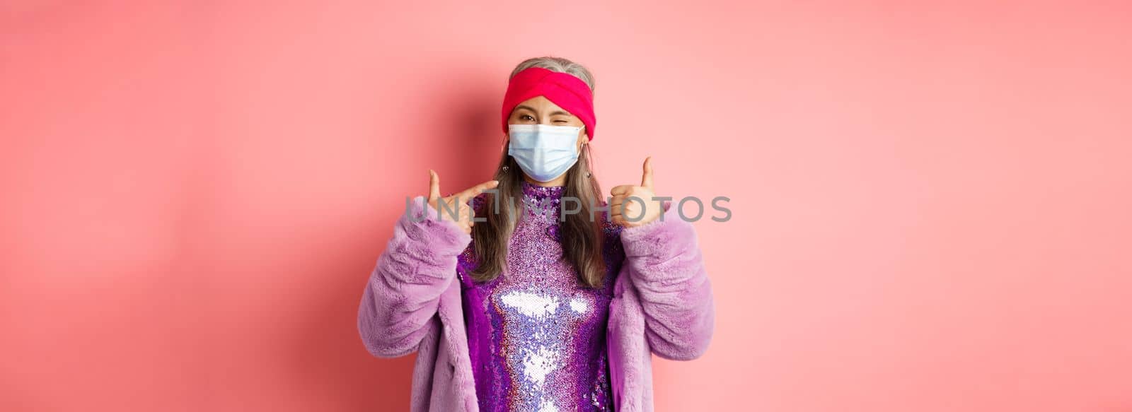 Covid-19, social distancing and fashion concept. Smiling asian senior woman in fashionable clothes and respirator, pointing at face mask and show thumb-up, pink background.
