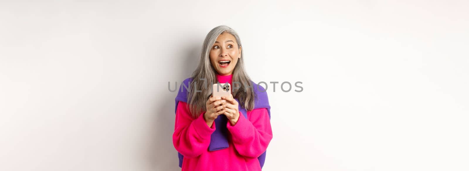 Amazed asian woman smiling and looking aside fascinated after reading promotion on smartphone, standing with mobile phone over white background.