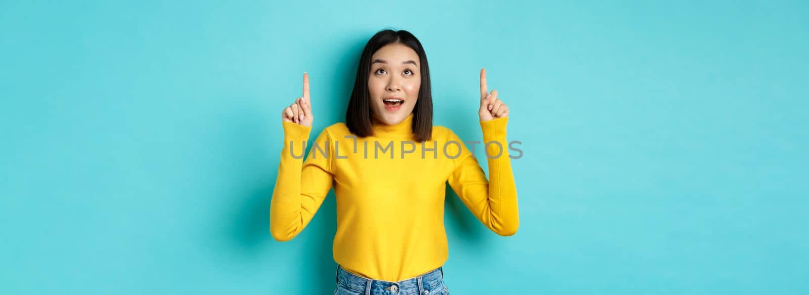 Shopping concept. Dreamy korean woman looking with desire at promo offer, pointing fingers up and staring at advertisement amazed, standing over blue background.