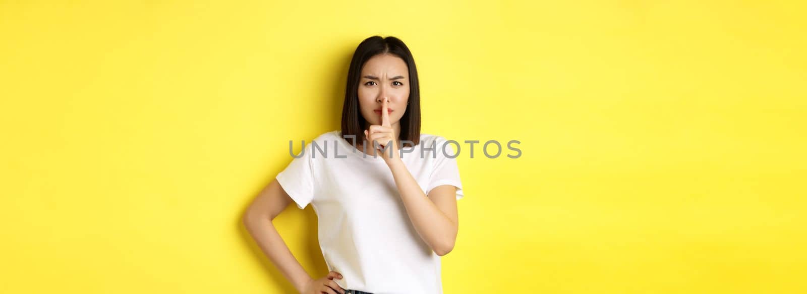 Beauty and fashion concept. Angry and bossy asian girl telling to be quiet, scolding loud people, shushing and frowning at camera, standing in t-shirt over yellow background by Benzoix
