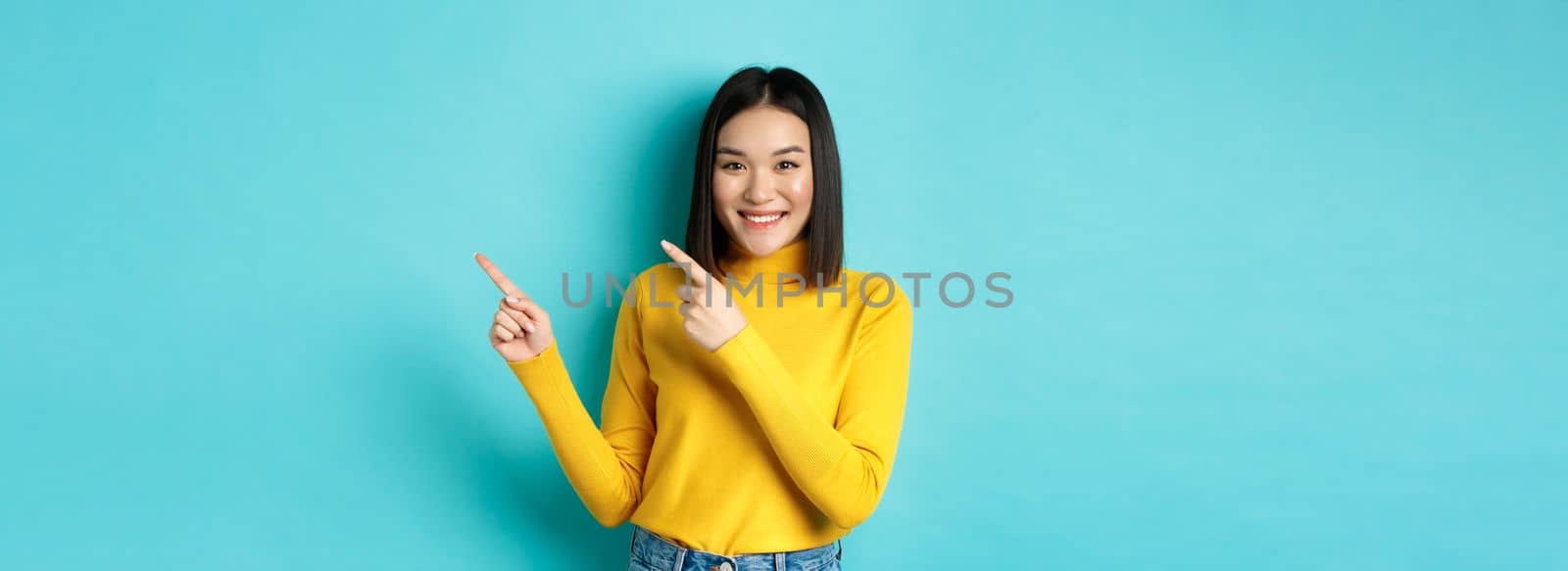 Shopping concept. Attractive asian girl in trendy sweater showing advertisement, pointing fingers right and smiling, recommend promotion deal, blue background by Benzoix